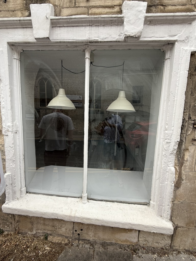 A building made of stone with a window featuring two light fixtures on either side, viewed through the glass. A man in a g...