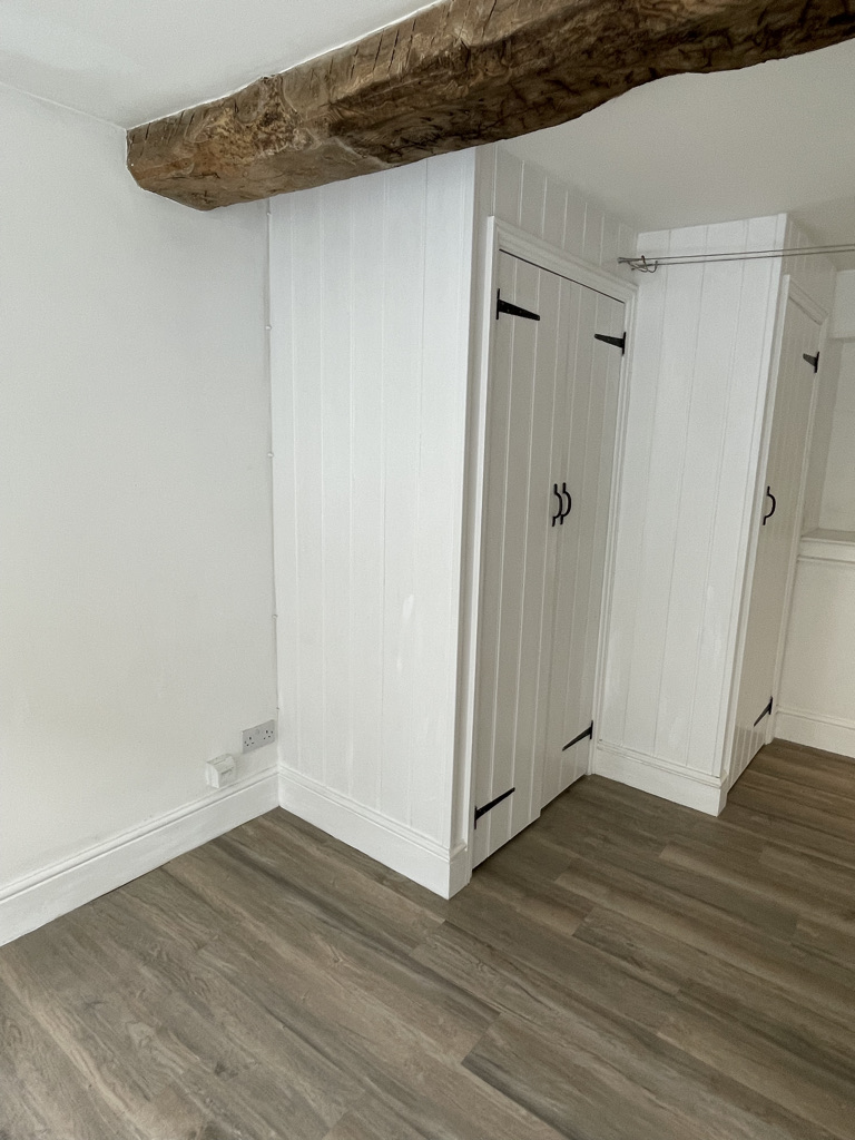 A vacant room with white paneled walls and grey laminated wooden floors. The ceiling has an exposed brown wooden beam, and...