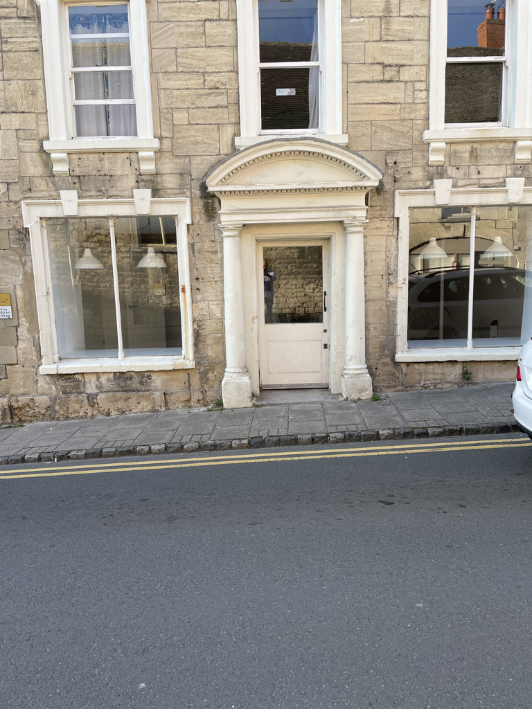 A cream-colored stone building with a white door and window frames, situated on a street with a white car parked inside th...